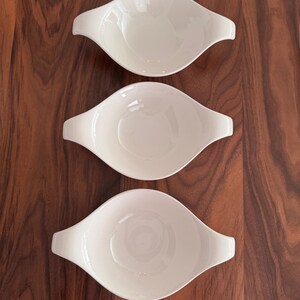 May include: Three white, oval-shaped serving dishes with handles are arranged on a dark wood surface. The dishes have a smooth, glossy finish and a simple, elegant design. These dishes are ideal for serving appetizers or small portions.