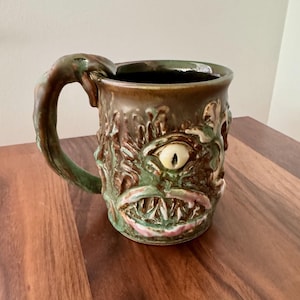 May include: A ceramic mug with a monster face design. The mug features a large, central eye, a wide mouth with sharp teeth, and a textured, green and brown exterior. The handle is shaped like a claw.