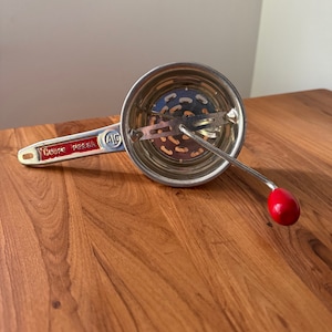 May include: A silver food mill with a red handle. The handle has the words "Coupe Persil" printed on it in red.