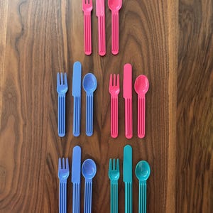May include: A collection of colorful plastic cutlery sets, including forks, knives, and spoons. The sets are in shades of pink, blue, and teal, arranged on a wooden surface. The utensils have a ribbed design.