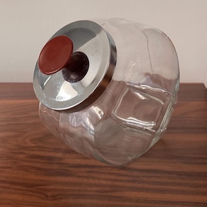 May include: A clear glass cookie jar with a stainless steel lid and a brown knob. The jar has a rounded shape with a square base and is sitting on a wooden surface.