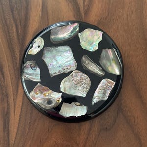 May include: A round, black coaster inlaid with iridescent abalone shell pieces. The shells display a range of colors, including pink, green, and silver, creating a decorative and unique design. The coaster sits on a dark wood surface.