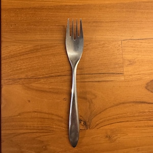 May include: A silver fork with four tines and a long, slender handle. The fork is lying flat on a wooden surface.