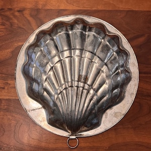 May include: A vintage, silver-toned metal mold in the shape of a seashell. The mold has a scalloped edge and a ring for hanging. The interior features detailed ridges, creating a textured surface. The antique kitchenware is likely used for baking or molding food.