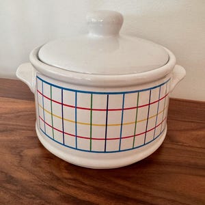 May include: A white ceramic casserole dish with a lid and two side handles. The dish features a colorful grid pattern in blue, red, yellow, and green. The casserole dish is sitting on a wooden surface.