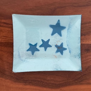 May include: A square, light blue dish with four dark blue stars. The dish has a slightly textured surface and is set against a dark wood background. The stars are arranged in a scattered pattern.