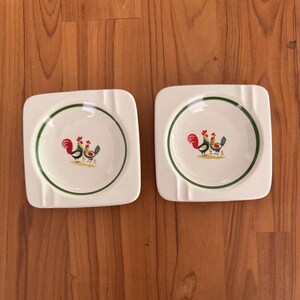 May include: Two square, white ceramic ashtrays with a green border and a rooster design. The rooster illustration features red, yellow, and green colors. The ashtrays are placed on a wooden surface.