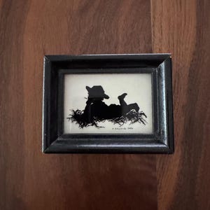 May include: A small rectangular black frame containing a silhouette of a person wearing a hat and lying on grass. The artwork is on a cream background and features the signature and date E. Edwards 1986 printed in the bottom right corner.