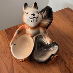 May include: A ceramic squirrel figurine with a bowl-shaped opening. The squirrel is painted in shades of brown, beige, and black, with a smiling face and a bushy tail. The figurine is designed to hold small items.