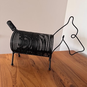 May include: Black metal wire dog sculpture. The dog is standing on a wooden surface. The dog's body is made of a coiled wire.
