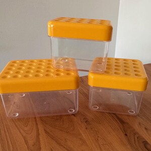 May include: Three clear plastic containers with yellow lids. The lids have multiple holes. The containers are stacked on a wooden surface. The containers are different sizes.