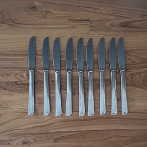 May include: Ten silver dinner knives laid out in a row on a wooden surface.
