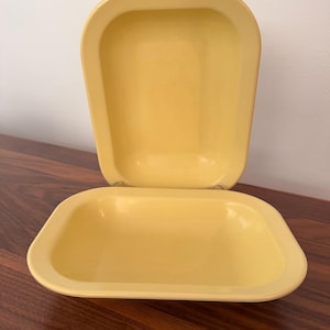 May include: Two rectangular yellow baking dishes with rounded corners and smooth, wide rims. One dish is positioned horizontally in the foreground, while the second dish is placed vertically behind it, showing the interior basin and rim.
