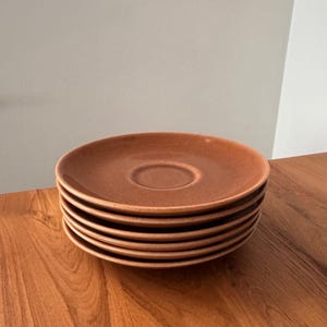 May include: A stack of six terracotta-colored ceramic saucers. Each saucer has a slightly raised center and a smooth, rounded edge. The saucers are stacked neatly on a wooden surface, with a neutral background.