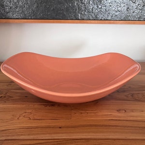 May include: A large, coral-colored ceramic serving bowl with a slightly curved, rectangular shape. The bowl is sitting on a wooden surface.