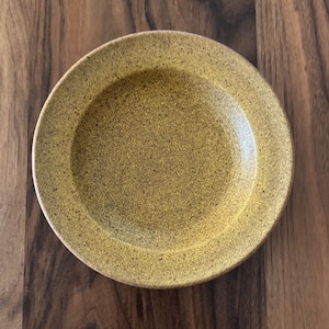 May include: A round, speckled yellow plate with a slightly raised rim. The plate has a textured surface and a warm, earthy tone. The plate is made of ceramic and is suitable for serving food.