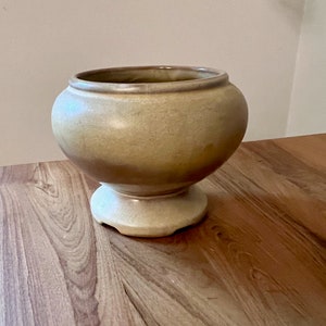 May include: A light brown ceramic planter with a wide, rounded base and a flared lip. The planter has a smooth, glazed finish and a subtle swirl pattern.