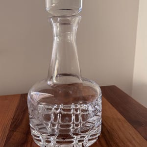 May include: A clear glass decanter with a stopper. The decanter has a bulbous base with a textured, patterned design. The neck is slender, and the stopper is a rounded shape. The decanter is sitting on a wooden surface.