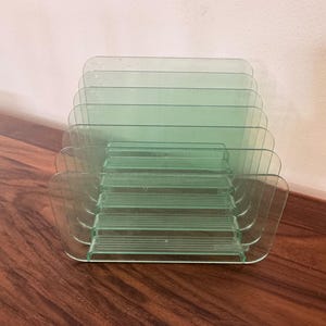 May include: A clear, green-tinted acrylic desk organizer with multiple slots. The organizer has a rectangular shape with rounded corners and a series of horizontal dividers. The design is simple and functional, suitable for office or home use.