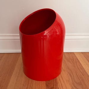 May include: A glossy red wastebasket with a cylindrical shape and a slanted top opening. The wastebasket is made of a smooth, reflective material and stands on a wooden floor. The interior is also red.