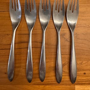 May include: Five silver forks with a simple, modern design. The forks are arranged in a row on a wooden surface.