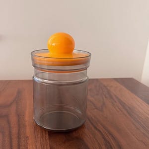 May include: A clear glass jar with an orange lid and a round orange knob. The jar is empty and sits on a dark wooden surface. The lid is flat with a circular design. The jar is suitable for storing dry goods.