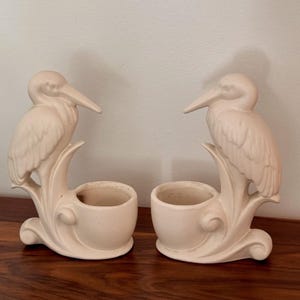 May include: A pair of matte off-white ceramic planters, each featuring a sculpted heron bird perched atop a curved base with an integrated circular bowl for holding small plants or decorative items, finished in a smooth, solid neutral tone.