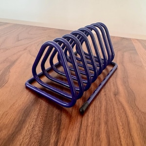 May include: A dark blue metal desk organizer with a series of triangular compartments. A matching dark blue pencil rests beside the organizer. The organizer is on a wooden surface.