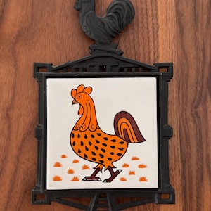 May include: Black metal trivet with a ceramic tile featuring an orange and brown rooster design. The trivet has a decorative rooster on top. The rooster on the tile has a dotted body and a brown tail. The tile is surrounded by a black metal frame.