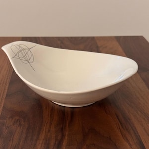 May include: Off-white, mid-century modern style serving bowl. The bowl has a unique, asymmetrical shape with a wide opening and a narrow base. A gray abstract design is on the side. The bowl is made of ceramic.