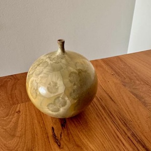 May include: A round, light brown ceramic vase with a white and light brown marbled pattern. The vase is sitting on a wooden surface.