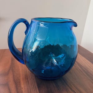 May include: A vibrant, hand-blown glass pitcher in shades of blue. The pitcher features a rounded body, a curved handle, and a pouring spout. The glass has a textured appearance, adding depth and visual interest to the piece. This decorative pitcher is ideal for serving beverages.