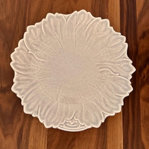 May include: A light-colored, leaf-shaped ceramic plate. The plate has a detailed vein pattern and a scalloped edge. The plate is set against a wooden background. The plate is likely used for serving food.