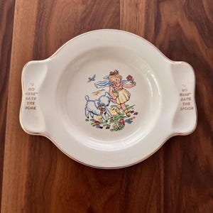 May include: Cream-colored vintage child's plate with a gold rim and handles. The plate features a colorful illustration of a girl with a lamb, flowers, and a bird. Text on the plate reads "I GO HERE SAYS THE FORK" and "I GO HERE SAYS THE SPOON."