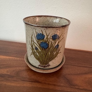May include: A speckled beige ceramic planter with a matching saucer, featuring a hand-painted illustration of blue thistle-like flowers with green stems and leaves. The pot has a dark brown rim and a cylindrical shape with a slightly flared top edge.