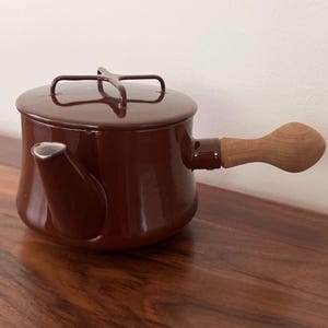 May include: A dark brown enameled metal teapot with a matching lid featuring a cross-shaped handle. The teapot has a short spout and a light-colored wooden handle extending from the side, providing a contrast to the smooth, glossy brown finish.