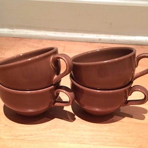May include: Four brown ceramic coffee cups are stacked in pairs. Each cup has a rounded bowl shape with a curved handle. The cups are a solid, warm brown color and appear to be made of stoneware.