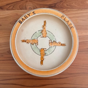 May include: A ceramic baby plate with an orange rim and the words "BABY'S PLATE" in black. The plate features four orange bunnies around a green circle. The plate is on a wooden surface.