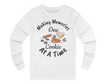 May include: White long-sleeve shirt with the text "Making Memories One Cookie At A Time" in black script. The graphic includes rolling pins, cookie cutters, and a mixing bowl, all in a cartoon style.