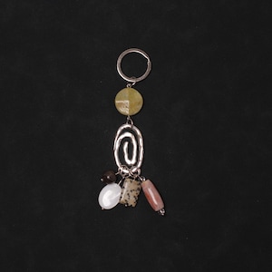 May include: A silver keyring with a decorative charm. The charm includes a green circular stone, a silver spiral, and small, colourful stones in white, brown, and pink. The background is black.