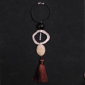 May include: A necklace featuring a black twisted cord, a smooth black bead, a hammered silver-tone irregular ring, a vertical strand of small white pearls, a large oval cream-colored stone, and a dark brown silk tassel hanging at the bottom.