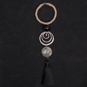 May include: A necklace featuring a beige twisted rope cord, a large black spherical bead, three concentric silver-toned rings, a single small white pearl, a circular grey marbled stone pendant, and a long black fabric tassel at the bottom.