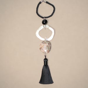 May include: A pendant necklace with a black rope loop, a black bead, a silver irregular ring, a patterned stone, and a black tassel. The necklace is designed to be worn as a pendant.