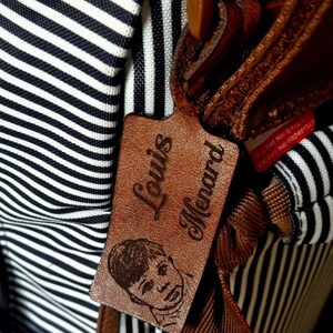 Puede incluir: Primer plano de una etiqueta de cuero marrón con el nombre "Louis Menard" en relieve en fuente cursiva. Una pequeña ilustración de una persona está debajo del nombre. La etiqueta está sujeta a una bolsa con tela a rayas blancas y negras.