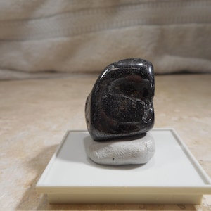 Puede incluir: Una piedra preciosa negra pulida, con una superficie brillante, colocada sobre una pequeña piedra gris. Las piedras se exhiben sobre una plataforma rectangular blanca, sobre un fondo neutro. La forma de la piedra es irregular y orgánica.