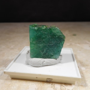 May include: A vibrant, emerald-green mineral specimen. The rough-cut gemstone is mounted on a white rectangular base with a gray adhesive. The stone's surface has visible texture and variations in color, showcasing its natural beauty.