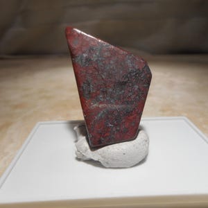 May include: A polished, irregularly shaped piece of red and dark gray jasper stone. The stone features a mottled, speckled texture with deep crimson and charcoal-colored inclusions throughout its surface, displayed upright on a small white base.