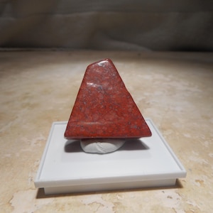 May include: A polished, trapezoidal-shaped red stone with speckled grey and white inclusions. The stone is mounted on a small, white, square display stand, which is supported by a white adhesive putty base to keep it upright for viewing.