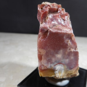 Puede incluir: Un espécimen de piedra en bruto de color marrón rojizo con una apariencia en capas. La piedra tiene una mezcla de colores, incluyendo rojos intensos, marrones y toques de amarillo. La roca se exhibe sobre un soporte negro, mostrando su textura natural.