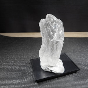 Puede incluir: Un cristal transparente y translúcido con una superficie rugosa e irregular. El cristal se muestra sobre una base cuadrada negra.
